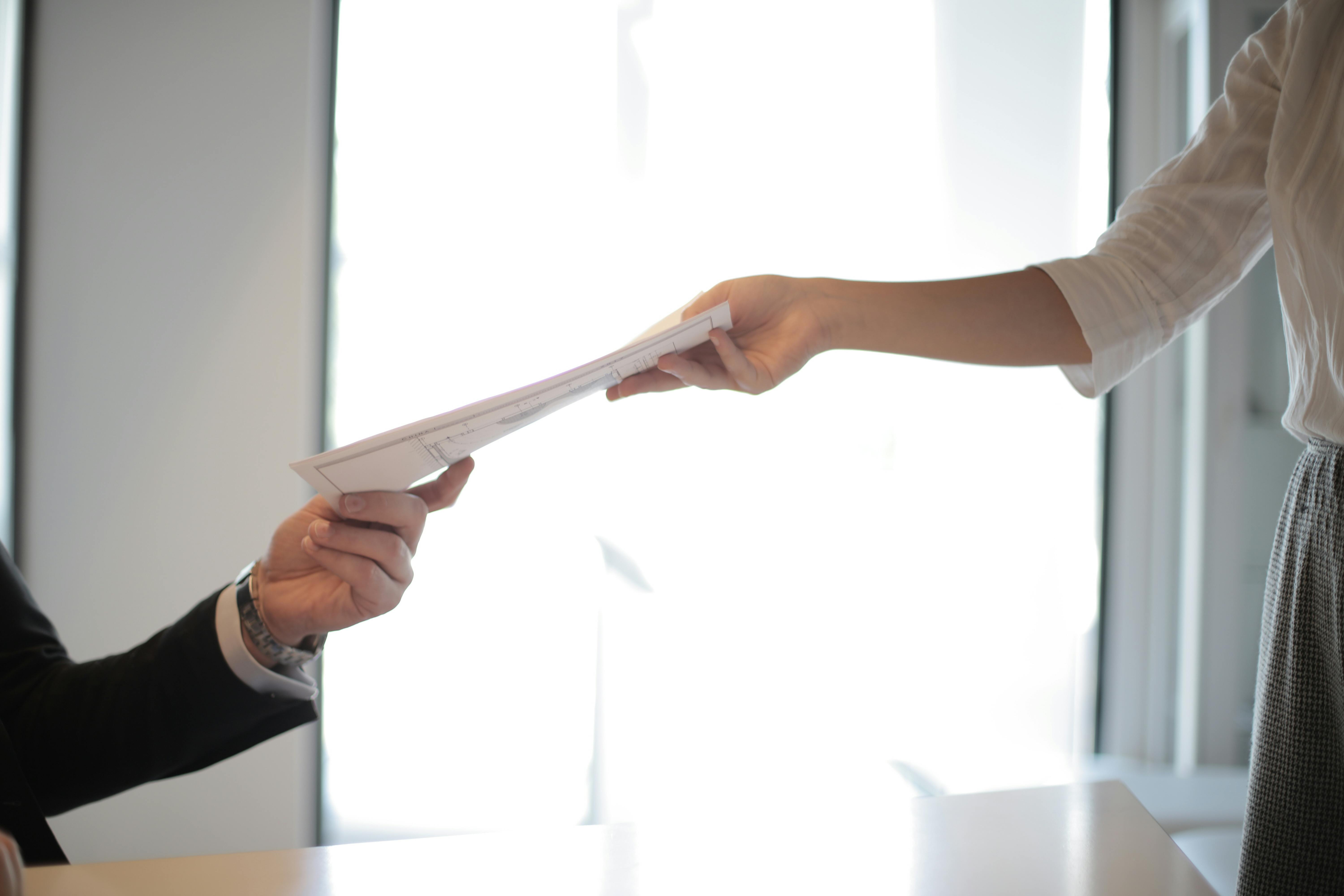 A person is handing over a document to another person, who is reaching out to receive it. The image captures the transaction from a close-up perspective, focusing on the hands and the document. The background is bright and softly lit, suggesting a professional or office environment.