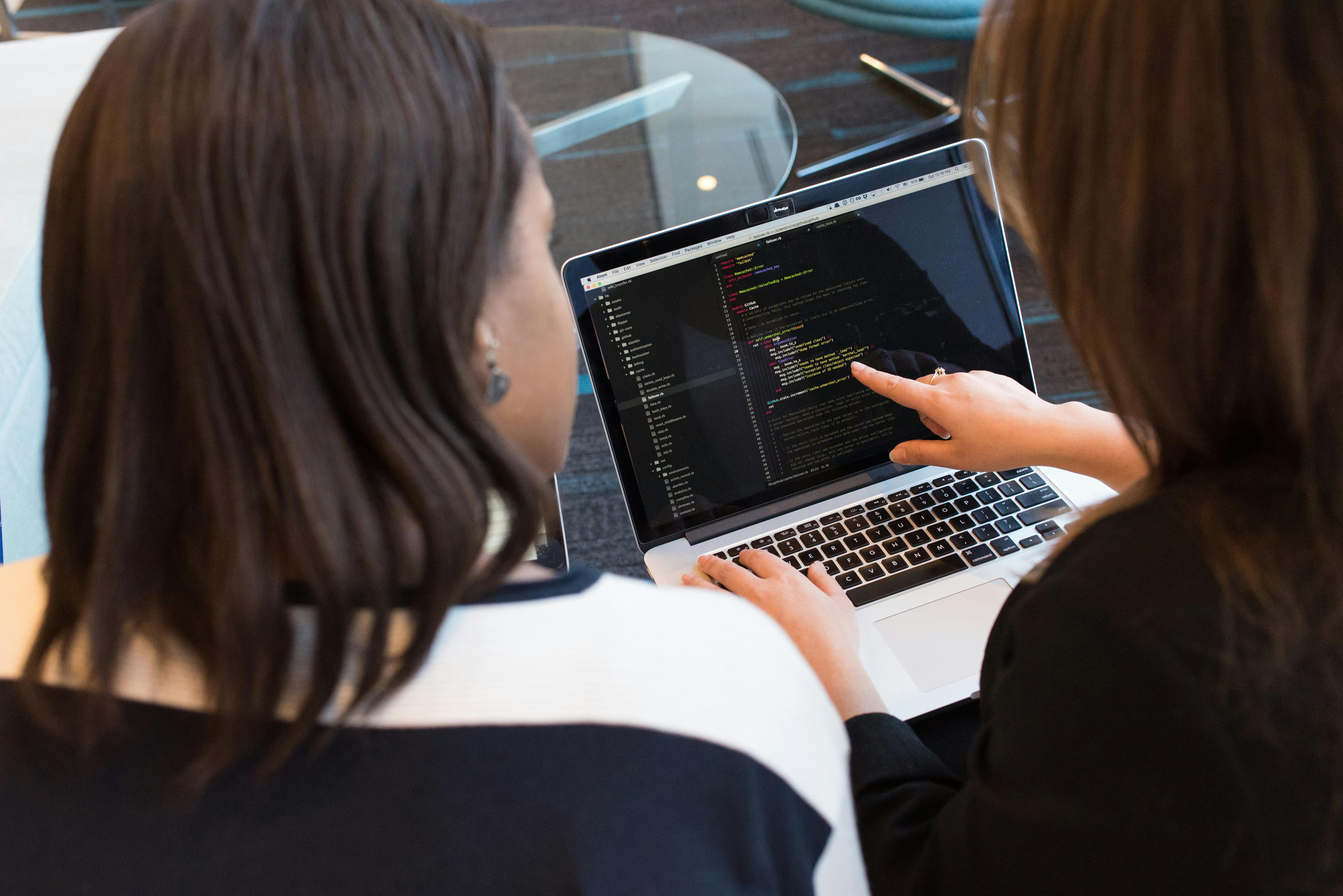 Two women looking at code at the laptop while one of the women is pointing to a specific line of code